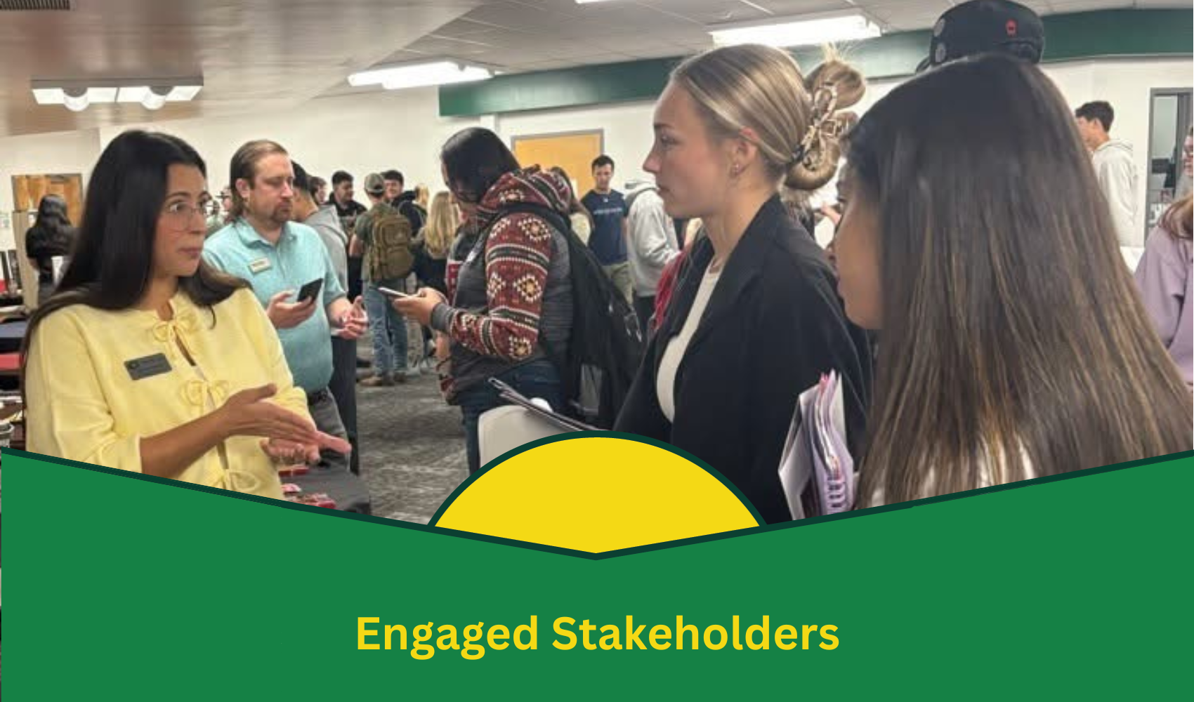 The image shows a busy indoor setting where several people are engaged in conversation. On the left, a woman in a yellow blouse is gesturing with her hands while speaking to two young women on the right. One of the young women has a notebook in hand, listening attentively. Other people are visible in the background, some interacting with each other and others using their phones. The environment appears to be a meeting or networking event, with fluorescent lighting and a casual atmosphere. At the bottom of the image, a green and yellow graphic design with the text "Engaged Stakeholders" is prominently displayed.